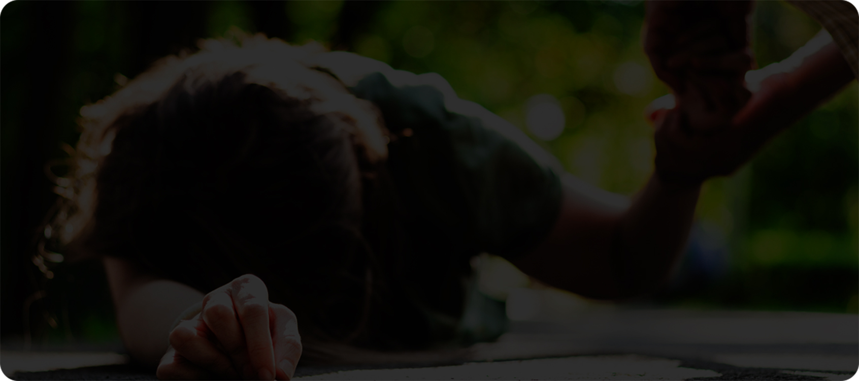 woman lying on the ground face first, resting on her right forearm. Someone above her is holding her left hand and appears to be helping her